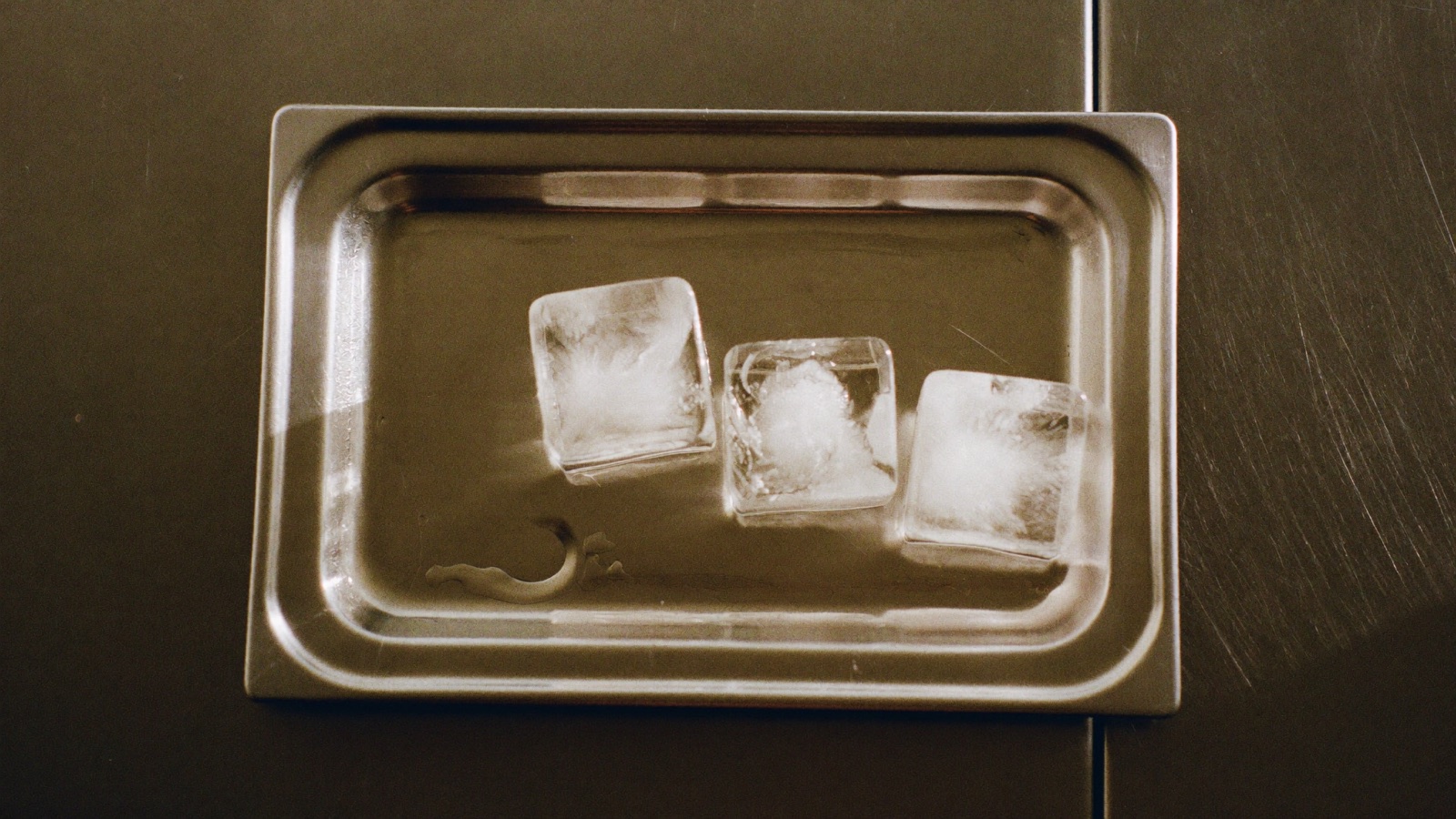 A stainless steel ice cube tray on a dark counter with three ice cubes left in it, meltwater pooling around them.