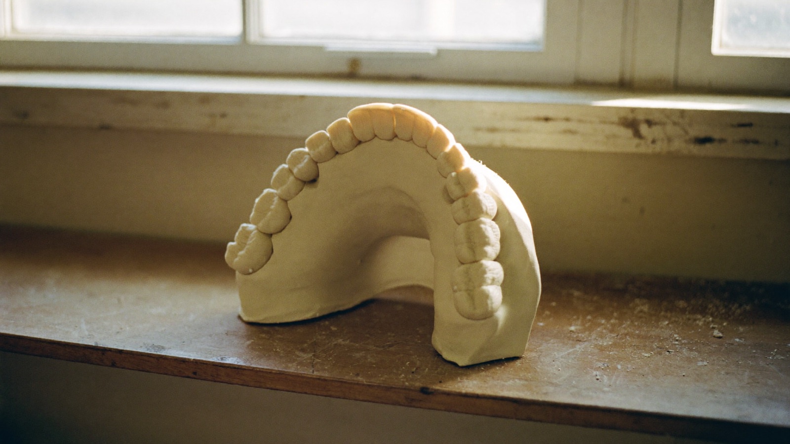 A plaster dental model of an upper arch sitting on a worn wooden laboratory bench beside a window.