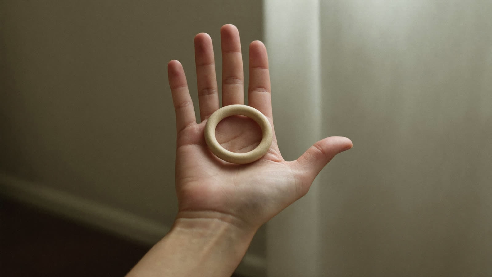 An adult's open hand holding a smooth wooden teething ring, in soft morning light.
