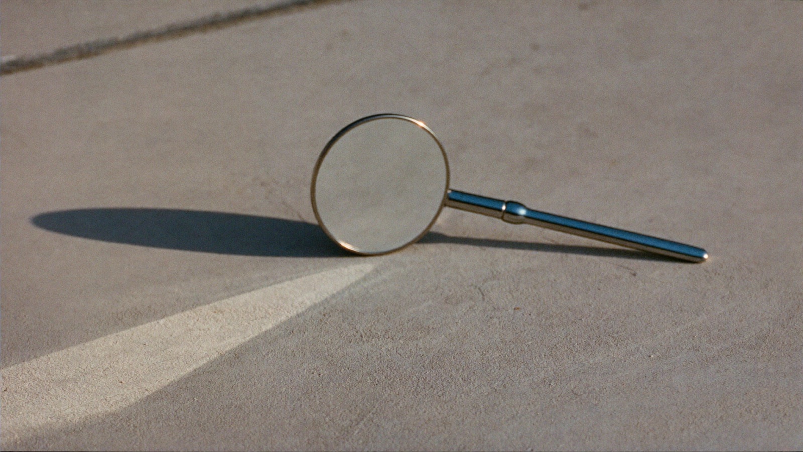 A small round dental mirror standing on a pale concrete surface in hard sunlight, casting a long shadow.