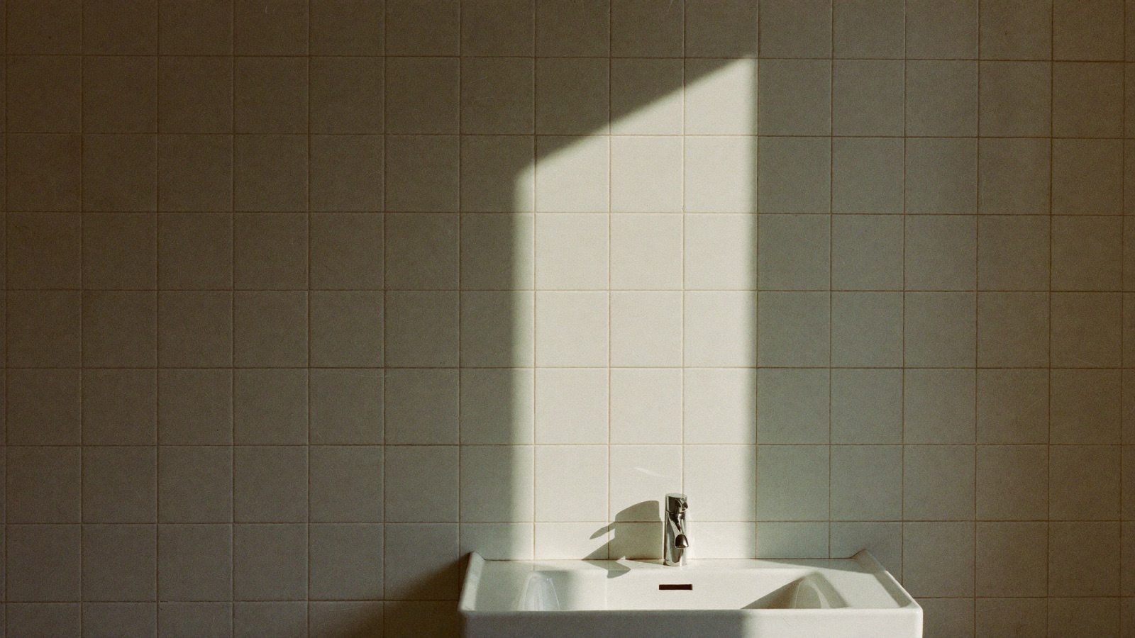 A white subway tiled bathroom wall with a chrome tap over a basin and a hard rectangle of morning sunlight across the tile.