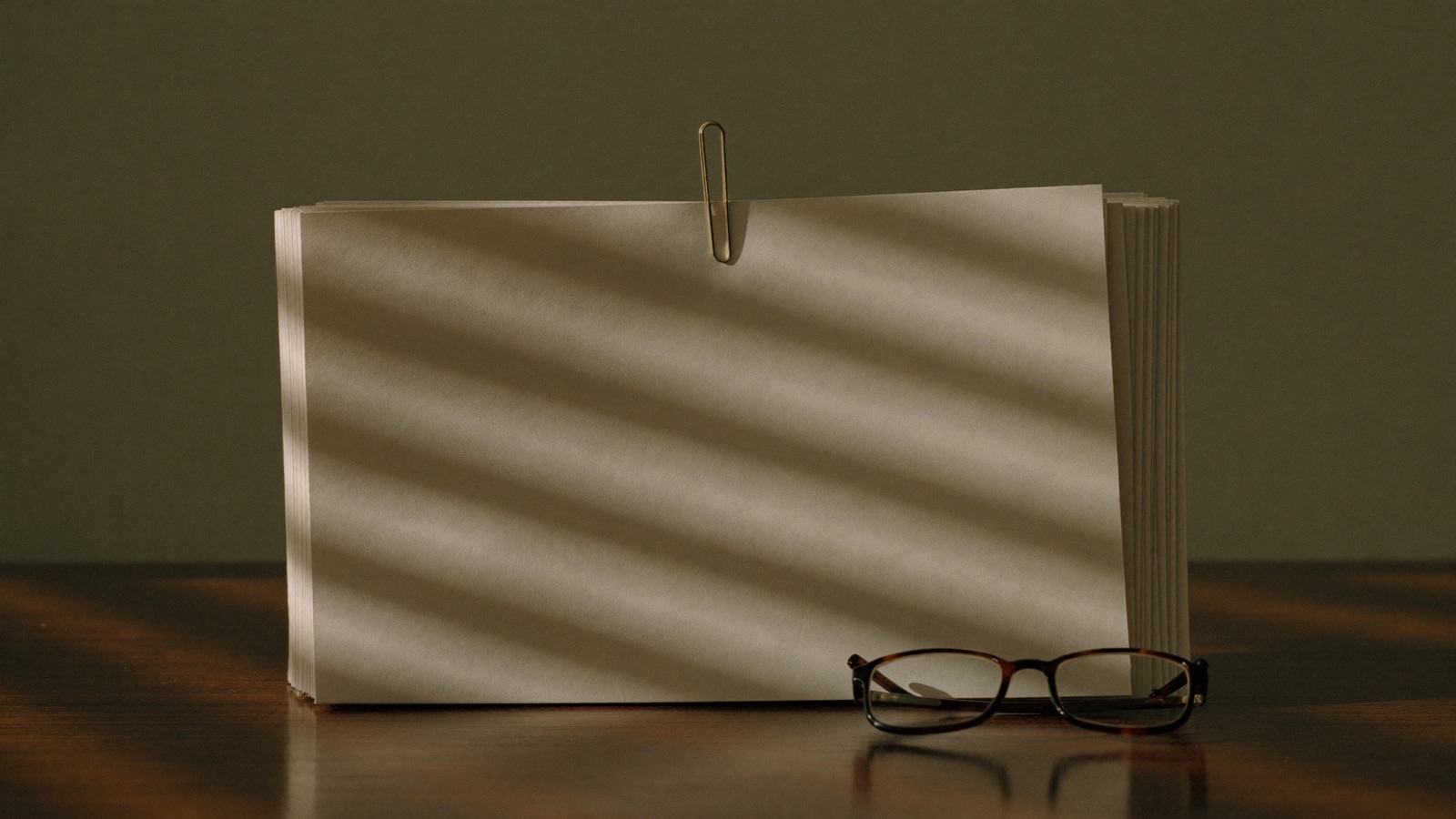 A stack of blank unprinted paper held by a paperclip on a desk, striped with light through window blinds, next to a pair of reading glasses.