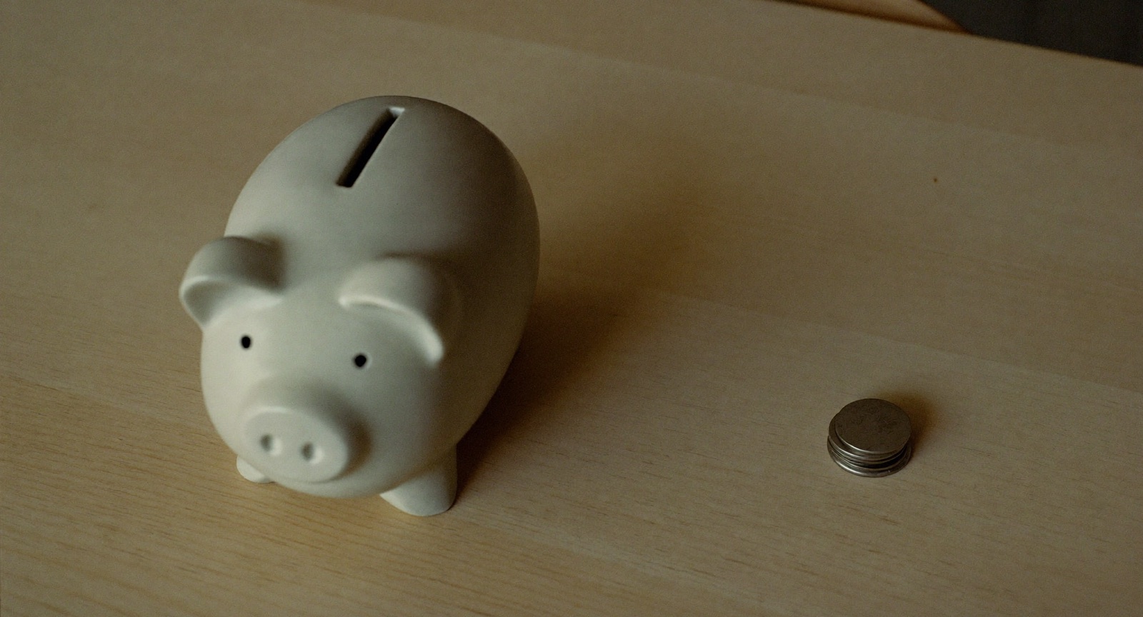 A plain white ceramic piggy bank on a pale wooden table beside a small stack of coins.
