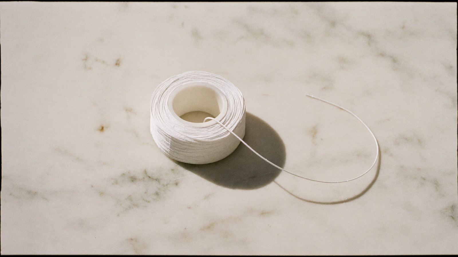 A spool of dental floss on a white marble surface with a single thread unspooled in a long curve.