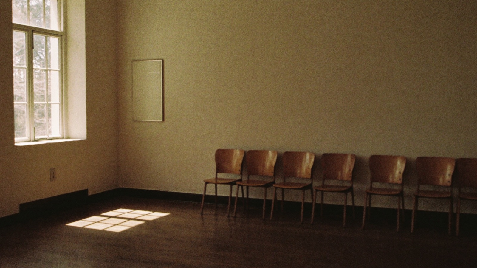 A row of empty wooden chairs along a bare wall in a waiting room, with afternoon light falling through a tall window onto the floor.