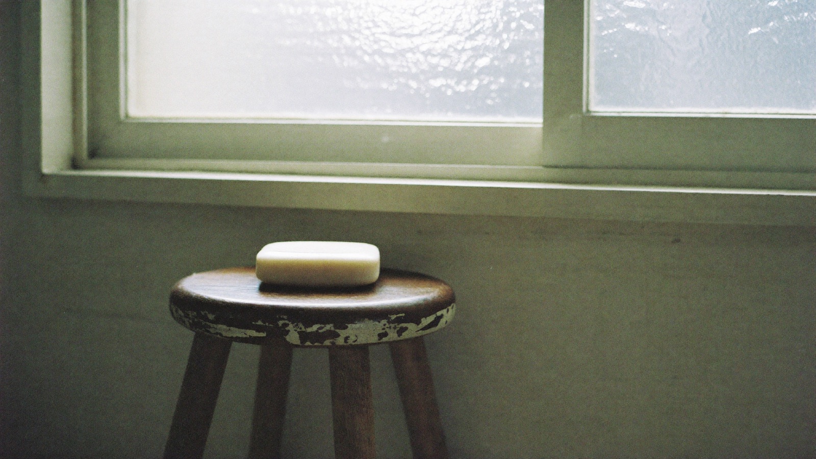 A plain white bar of soap on a small worn wooden stool below a window, lit by cool morning light.