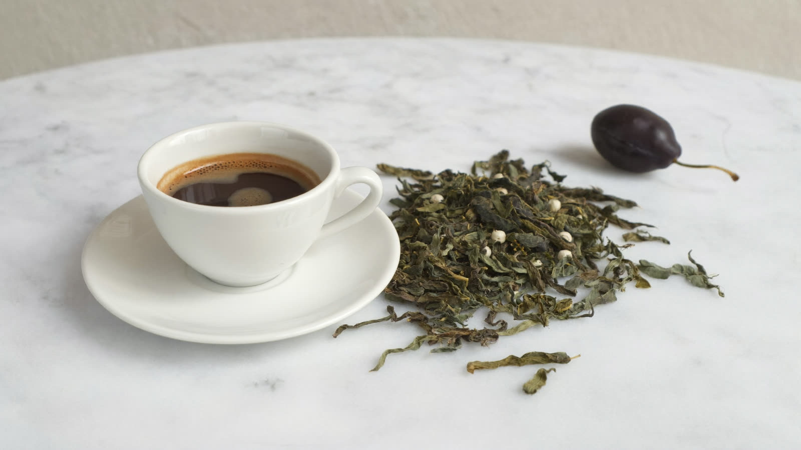 An espresso cup, loose tea leaves and a dark plum on a marble counter in bright daylight.