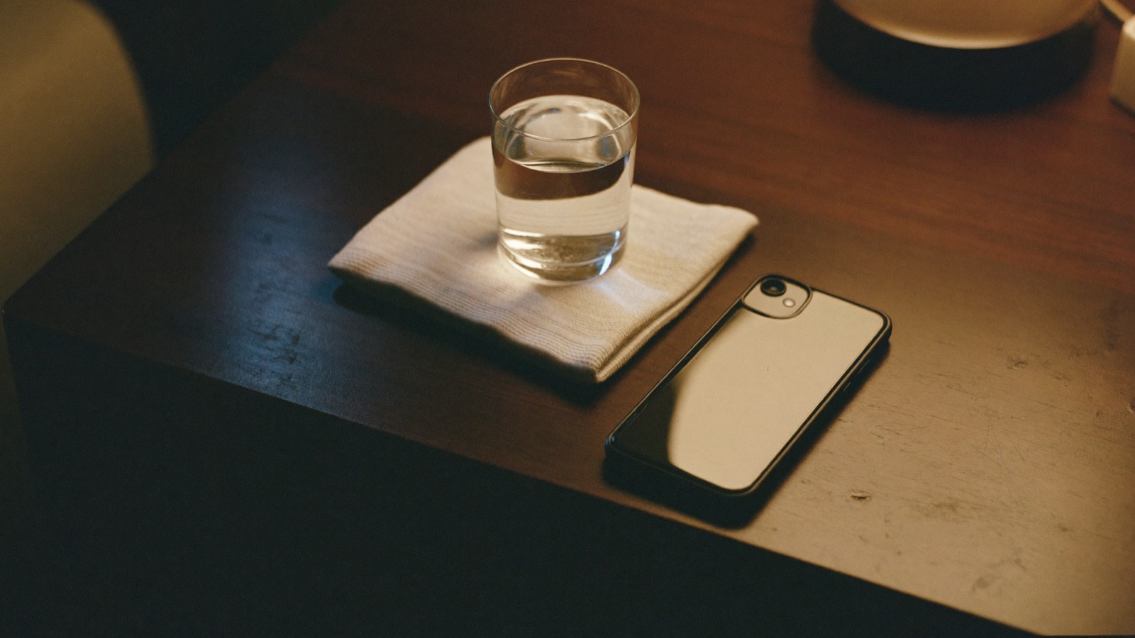 A glass of water and a folded washcloth on a dark bedside table at night, with a phone lying face down beside them under a warm lamp.